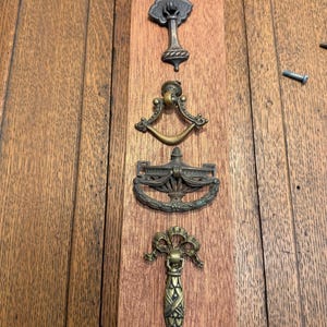 May include: Four antique bronze drawer pulls and a tree-shaped handle are arranged vertically on a wooden surface. The pulls have ornate designs, including a tree, a curved handle, a wreath, and a bow. The wood has a warm, brown tone.