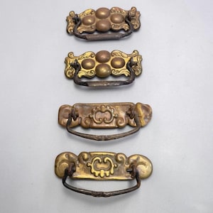 May include: Four metal drawer pulls arranged vertically. The top two feature a cluster of five raised circular domes with ornate edges. The bottom two feature a central scrollwork design on a rectangular plate. All have dark, curved drop handles.