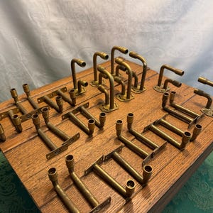May include: A collection of vintage brass hardware pieces, including brackets and curved pipe fittings, arranged on a wooden surface. The hardware has a warm, aged patina, suggesting antique or reclaimed origins. The pieces vary in size and shape, suitable for various home decor projects.