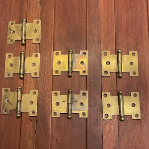 May include: Nine brass door hinges arranged on a wooden surface. Each hinge has a rectangular shape with rounded corners and multiple screw holes. The hinges are a warm, aged gold color.