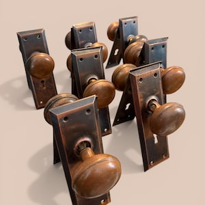 May include: A collection of antique door knobs and backplates. The knobs are a warm, copper color, and the backplates are a dark, aged bronze. The door hardware has a vintage aesthetic, with keyholes and mounting holes visible.