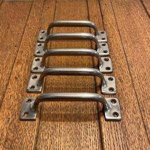 May include: Five silver-toned metal pull handles arranged in a vertical row. Each handle has a curved bar and two rectangular mounting plates with screw holes. The handles are set against a wooden surface.