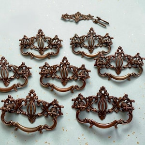 May include: A collection of ornate, bronze-colored drawer pulls and decorative hardware. The pulls feature intricate floral and scrollwork designs with curved handles. The hardware is arranged on a light blue surface.