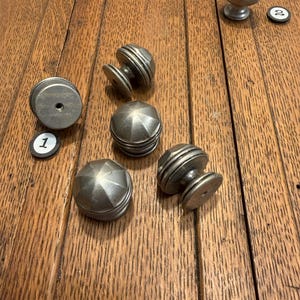 May include: A collection of antique metal cabinet knobs in various designs. The knobs are silver-toned and sit on a wooden surface. Some knobs have a round, ribbed design, while others have a faceted, star-like appearance. Two small numbered tags are also present.
