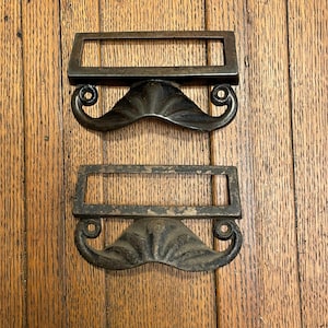 May include: Two antique bronze drawer pulls with a mustache design. Each pull features a rectangular frame above a stylized mustache with curled ends. The pulls are set against a wooden background.