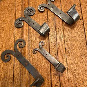 May include: Four wrought iron door latches with a rustic, aged appearance. Each latch features a decorative scroll design and a curved catch. The metal has a dark, textured finish, suggesting a handmade quality. These latches are suitable for doors or gates.