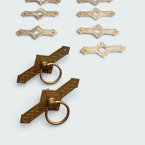 May include: A collection of antique brass hardware, including decorative diamond-shaped plates and door pulls with ring handles. The hardware has an aged, vintage appearance, with intricate etched patterns. The pieces are arranged on a white background.