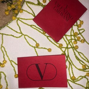 May include: Two red cards featuring the Valentino Garavani logo. One card displays the full brand name, while the other showcases the letter V within a circle.