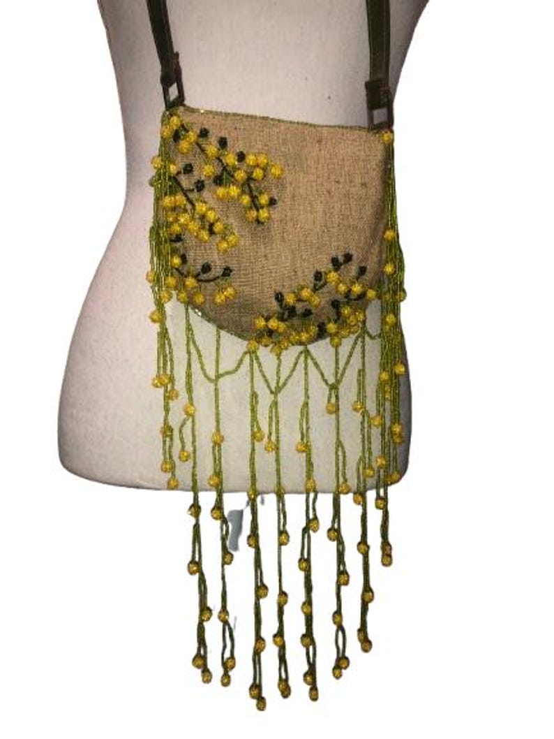 May include: A brown burlap crossbody bag with yellow and green beaded fringe. The bag features a floral design with yellow beads and black accents.