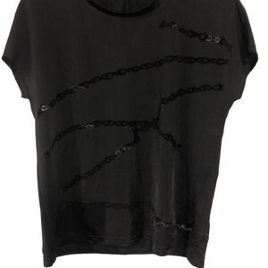 Può includere: T-shirt nero a maniche corte con scollo tondo e un motivo a catena decorativo sul davanti. La maglietta è appesa a una gruccia nera con il nome del marchio "Balenciaga" visibile.