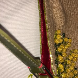 May include: A small, brown, burlap handbag with a red velvet trim and a green beaded strap. The bag is decorated with yellow and green beaded flowers.