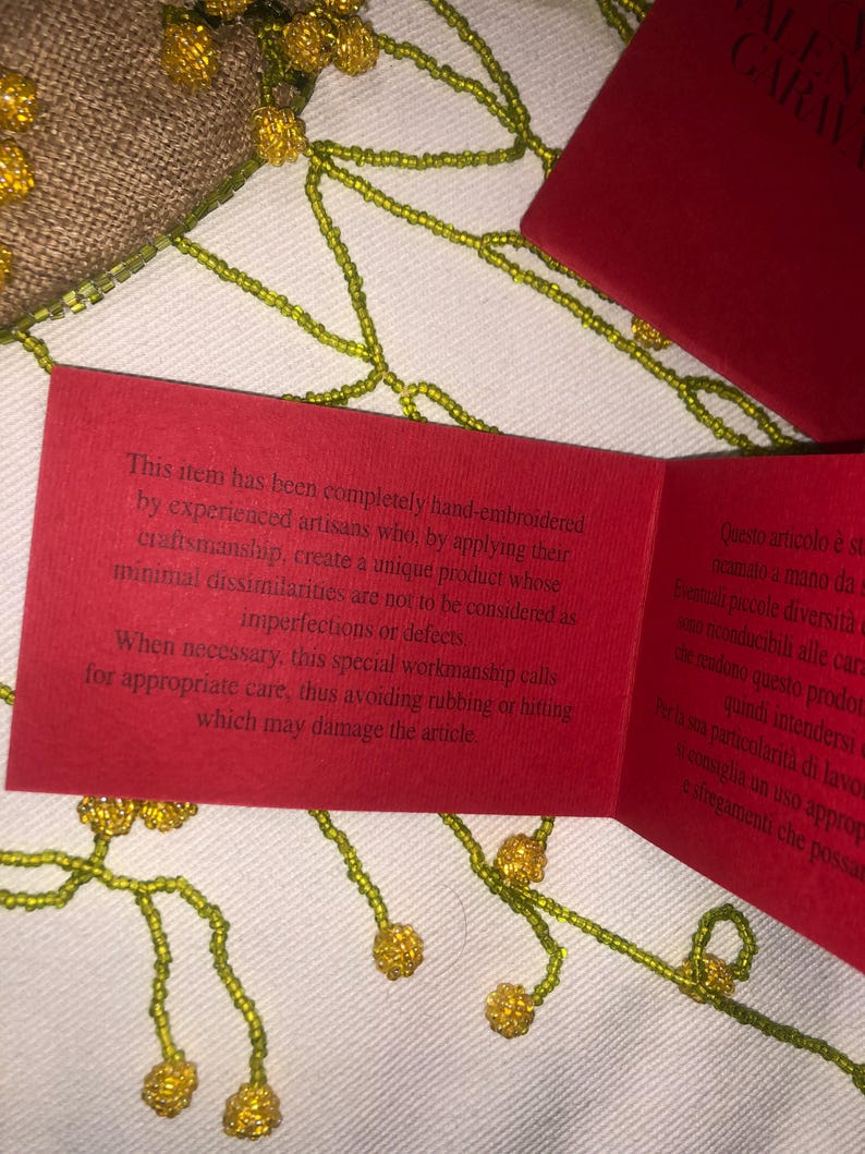 May include: A red card with white text explaining that the item is hand-embroidered and requires special care to avoid damage. The text reads: "This item has been completely hand-embroidered by experienced artisans who, by applying their craftsmanship, create a unique product whose minimal dissimilarities are not to be considered as imperfections or defects. When necessary, this special workmanship calls for appropriate care, thus avoiding rubbing or hitting which may damage the article."