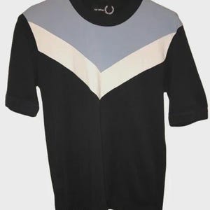 May include: A black short-sleeve t-shirt featuring a chevron-shaped color-blocked design across the chest in light blue and white. The shirt has a round neckline and includes the Raf Simons and Fred Perry laurel wreath logo on the inner collar.