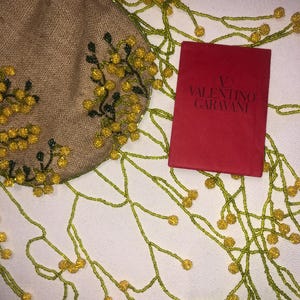 May include: A small, beige, beaded bag with yellow flowers and green leaves. The bag is attached to a long beaded chain with yellow beads and green beads. The bag is sitting on a white surface with a red box that says "VALENTINO GARAVANI" on it.