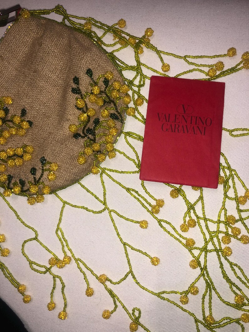 May include: A small, beige, beaded bag with yellow flowers and green leaves. The bag is attached to a long beaded chain with yellow beads and green beads. The bag is sitting on a white surface with a red box that says "VALENTINO GARAVANI" on it.