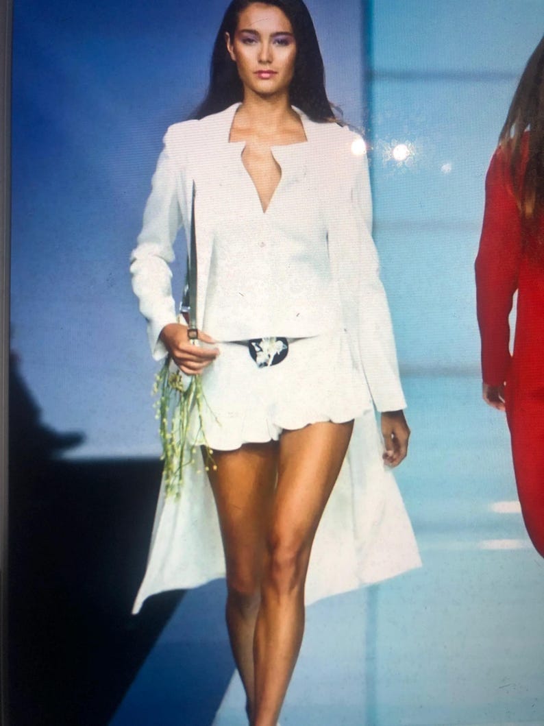 May include: A white long-sleeved jacket with a deep V-neckline and a matching white skirt. The model is wearing a black belt with a silver buckle.