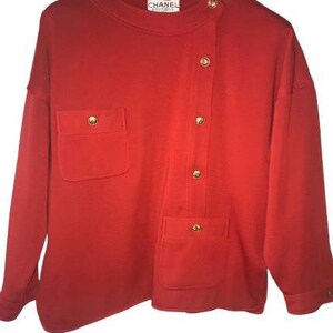Può includere: Una camicia rossa a maniche lunghe con chiusura a bottoni e una tasca sul lato sinistro. La camicia ha un'etichetta Chanel Boutique.
