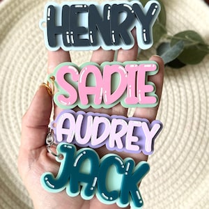 May include: Four custom name keychains featuring layered acrylic designs. The top tag is dark gray with light blue backing, the second is pink with light green, the third is light purple with white, and the bottom is dark teal with light blue.