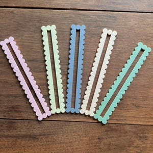 May include: Five pastel-colored, scalloped-edge, slotted craft sticks arranged on a wooden surface. Colors include light pink, green, blue, white, and mint green. The sticks have a decorative, wavy edge.