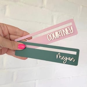 Può includere: Due segnalibri rettangolari con una fessura ritagliata nella parte superiore. Un segnalibro è rosa chiaro con il nome COURTNEY in lettere stampatello bianche. L'altro è verde scuro con il nome Megan in corsivo bianco.