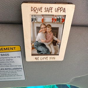 May include: A wooden car visor photo frame with the text "DRIVE SAFE UPPA" at the top and "WE LOVE YOU" at the bottom. The frame holds a photo of a woman and a child hugging in front of a fireplace.