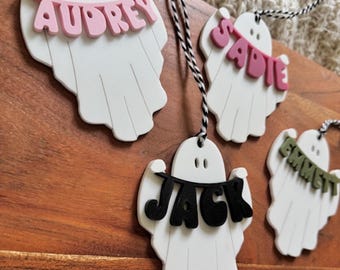 May include: White ghost-shaped decorations with names on banners. The names Audrey, Sadie, Jack, and Emmett are displayed in pink, black, and green lettering. Each decoration has a black and white string for hanging.