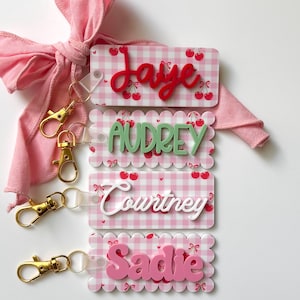 Op de afbeelding: Vier rechthoekige sleutelhangers met een roze en wit gingham-patroon met kleine rode kersen. Elke sleutelhanger toont een naam in reliëf: Jaye in rood, Audrey in saliegroen, Courtney in wit en Sadie in lichtroze.