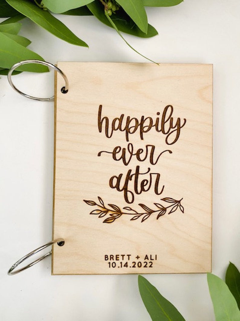 K&ouml;nnte beinhalten: Ein G&auml;stebuch aus Holz mit Ringbindung. Der Einband ist mit dem Text "happily ever after" und einem floralen Design graviert. Die Namen "Brett + Ali" und das Datum "10.14.2022" sind ebenfalls auf dem Einband graviert.