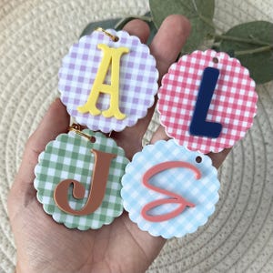 May include: Four circular scalloped-edge tags featuring gingham patterns in lavender, pink, sage green, and light blue. Each tag displays a raised letter in a contrasting color: a yellow A, a navy blue L, a terracotta J, and a coral pink S.