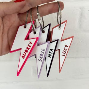 May include: A collection of white lightning bolt-shaped keychains with names Audrey, Sadie, Mia, and Lucy. Each keychain has a different colored border: pink, lavender, black, and coral. The keychains are attached to silver wire loops.