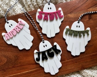 Könnte beinhalten: Vier weiße, geisterförmige Ornamente mit schwarz-weißen Schnuraufhängern. Jeder Geist hält ein Banner mit einem Namen: Audrey (rosa), Sadie (rosa), Jack (schwarz) und Emmett (grün). Die Ornamente befinden sich auf einer Holzoberfläche.