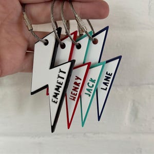 Könnte beinhalten: Eine Sammlung personalisierter Blitz-Schlüsselanhänger. Jeder Schlüsselanhänger hat die Form eines weißen Blitzes mit einem farbigen Rand und einem aufgedruckten Namen: Emmett, Henry, Jack und Lane. Die Schlüsselanhänger sind an silbernen Drahtringen befestigt.