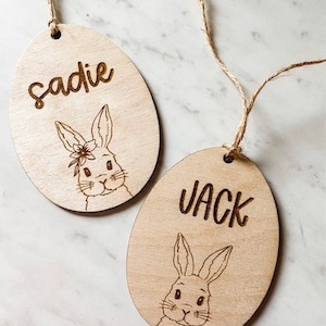Op de afbeelding: Twee houten paaseieren ornamenten met konijnen illustraties en de namen "Sadie" en "Jack" erop gegraveerd.