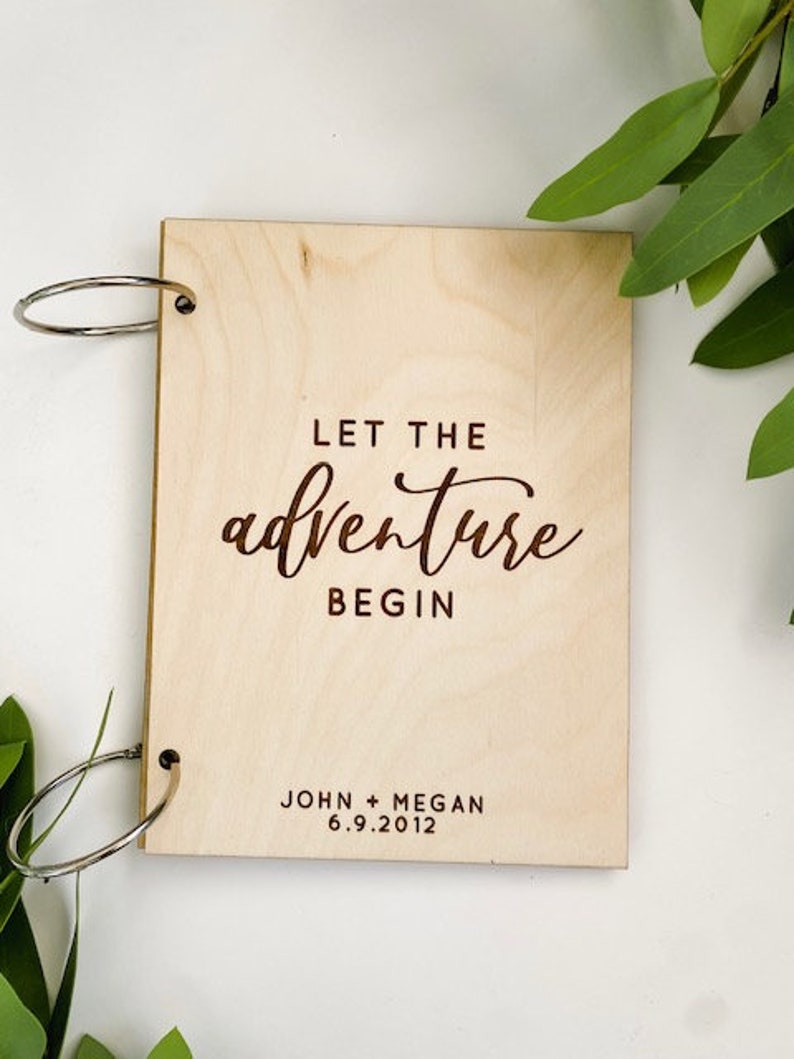 K&ouml;nnte beinhalten: Ein G&auml;stebuch aus Holz mit Ringbindung. Der Einband ist mit dem Text "Let the adventure begin" und den Namen "John + Megan" sowie dem Datum "6.9.2012" graviert.