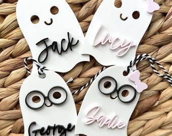 Op de afbeelding: Vier witte cadeau-etiketten in de vorm van een spook met zwarte en roze details. De etiketten zijn versierd met verschillende gezichten, waaronder een glimlachende spook, een spook met een bril en een spook met een strik. De etiketten zijn gelabeld met de namen "Jack", "Lucy", "George" en "Sadie".