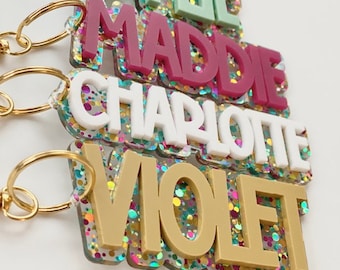 May include: Four colorful glitter keychain charms with the names "Kylie", "Maddie", "Charlotte", and "Violet" written in bold letters. Each charm is attached to a gold key ring.