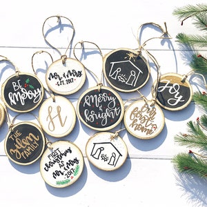 Peut inclure: Un ensemble de 10 décorations rondes en bois avec des motifs peints en noir et blanc. Les décorations présentent diverses phrases et images sur le thème de Noël, notamment "Be Merry", "Noel", "Joy", "Our First Home", "Merry & Bright", "The Zilen Family", "First Christmas as Mr. & Mrs. Est. 2017", et une scène de la nativité.