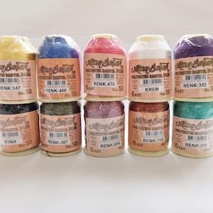Op de afbeelding: Tien spoelen garen in verschillende kleuren, waaronder goud, blauw, rood, wit, paars, zwart, groen, roze, oranje en teal. Elke spoel is gelabeld met de merknaam "Altın Başak" en de kleurcode. De spoelen zijn gerangschikt in twee rijen van vijf.