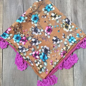 Può includere: Una sciarpa quadrata con un motivo floreale nei toni del blu, rosa e bianco su sfondo marrone. I bordi sono rifiniti con un pizzo all'uncinetto rosa acceso. La sciarpa è piegata e presentata su una superficie di legno.