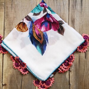 Può includere: Fazzoletto quadrato bianco con stampa floreale nei toni del viola, blu e marrone. I bordi sono rifiniti con un bordo all'uncinetto rosa e viola e un punto turchese. Esposto su una superficie di legno.