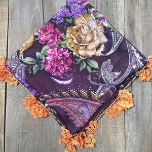 Può includere: Una sciarpa quadrata con un motivo floreale nei toni del viola, rosa e oro. La sciarpa ha uno sfondo viola scuro con un motivo paisley. I bordi sono rifiniti con pizzo arancione.