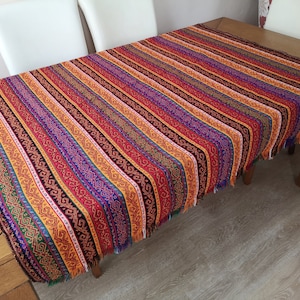 May include: A colorful striped table runner with a geometric pattern in shades of red, orange, yellow, green, blue, and purple. The runner has a fringe border and is made of woven fabric.