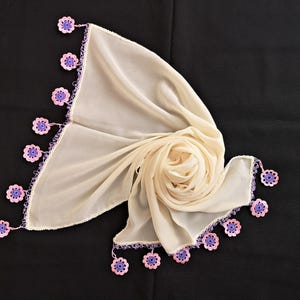 Può includere: Una sciarpa di colore crema trasparente con un bordo all'uncinetto con fiori rosa e viola.