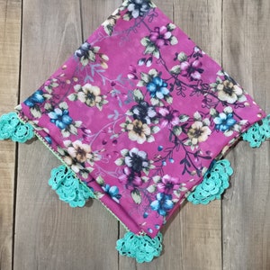 Può includere: Una sciarpa quadrata con motivo floreale nei toni del rosa, blu e bianco. La sciarpa presenta un motivo ripetuto di fiori e tralci. I bordi sono rifiniti con pizzo all'uncinetto turchese.