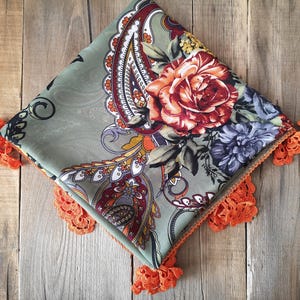 Può includere: Sciarpa quadrata con un design floreale e paisley. La sciarpa presenta una grande rosa arancione e fiori blu su uno sfondo verde salvia. I bordi sono rifiniti con pizzo arancione.