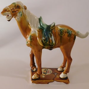 May include: A brown glazed ceramic horse figurine with green and white accents. The horse is wearing a saddle and has a long tail. The figurine is standing on a brown base with a white label that says "100% Hand Made".