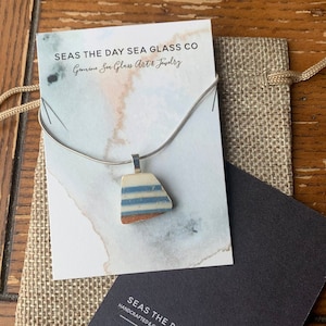 May include: A silver chain necklace with a pendant made from a piece of sea glass. The sea glass is blue and white with a brown edge. The pendant is hanging on a card that says "Seas the Day Sea Glass Co. Handmade & Genuine Sea Glass Jewellery by Cassie Kelly." The card also includes the website and social media handles for the company.
