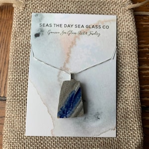 May include: A silver chain necklace with a pendant made from a piece of blue and gray sea glass. The necklace is displayed on a white card with the text "Seas the Day Sea Glass Co. Genuine Sea Glass Art & Jewelry".