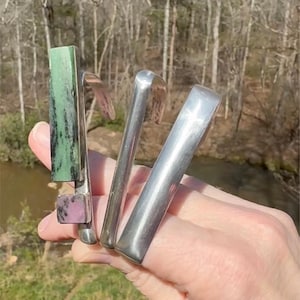May include: A silver-toned ring with three bands. One band features a rectangular green and black stone, and a square pink stone. The other two bands are smooth and curved. The ring is held up against a natural outdoor background.
