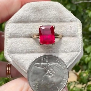 May include: A gold ring featuring a large, rectangular, faceted red gemstone set in a four-prong mounting. The ring is displayed on a light-colored, hexagonal velvet ring box next to a 2022 United States quarter coin for scale reference.